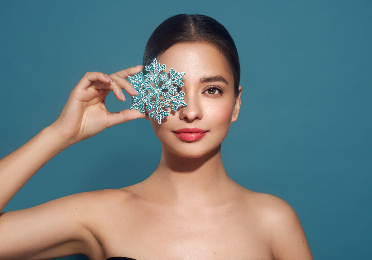 Beautiful young woman with red lipstick, shiny clean skin and face, showing glowing snowflake, near face and smiling, isolated on a blue background. Christmas holidays concept