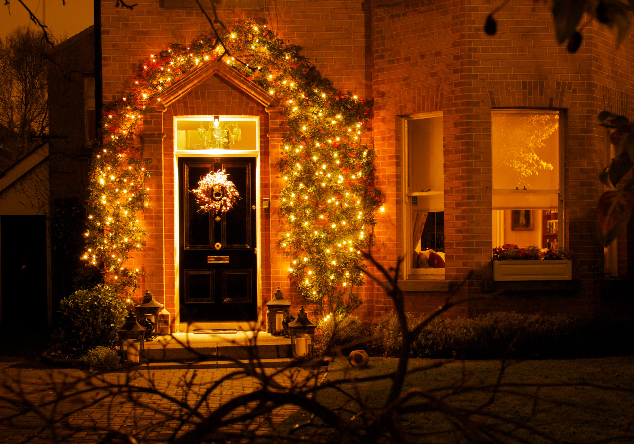 One traditional black Dublin door decorated with Christmas lights and two windows on birck wall at night, some branches at the front.