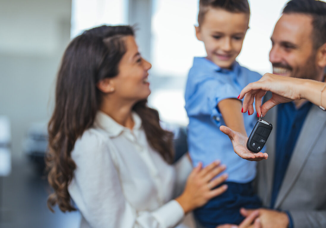 Happy parents with small kid after receiving keys for their new car in a showroom. Congratulations, we have a deal about buying a car! Happy family came to an agreement with a car salesperson at a meeting in a showroom.