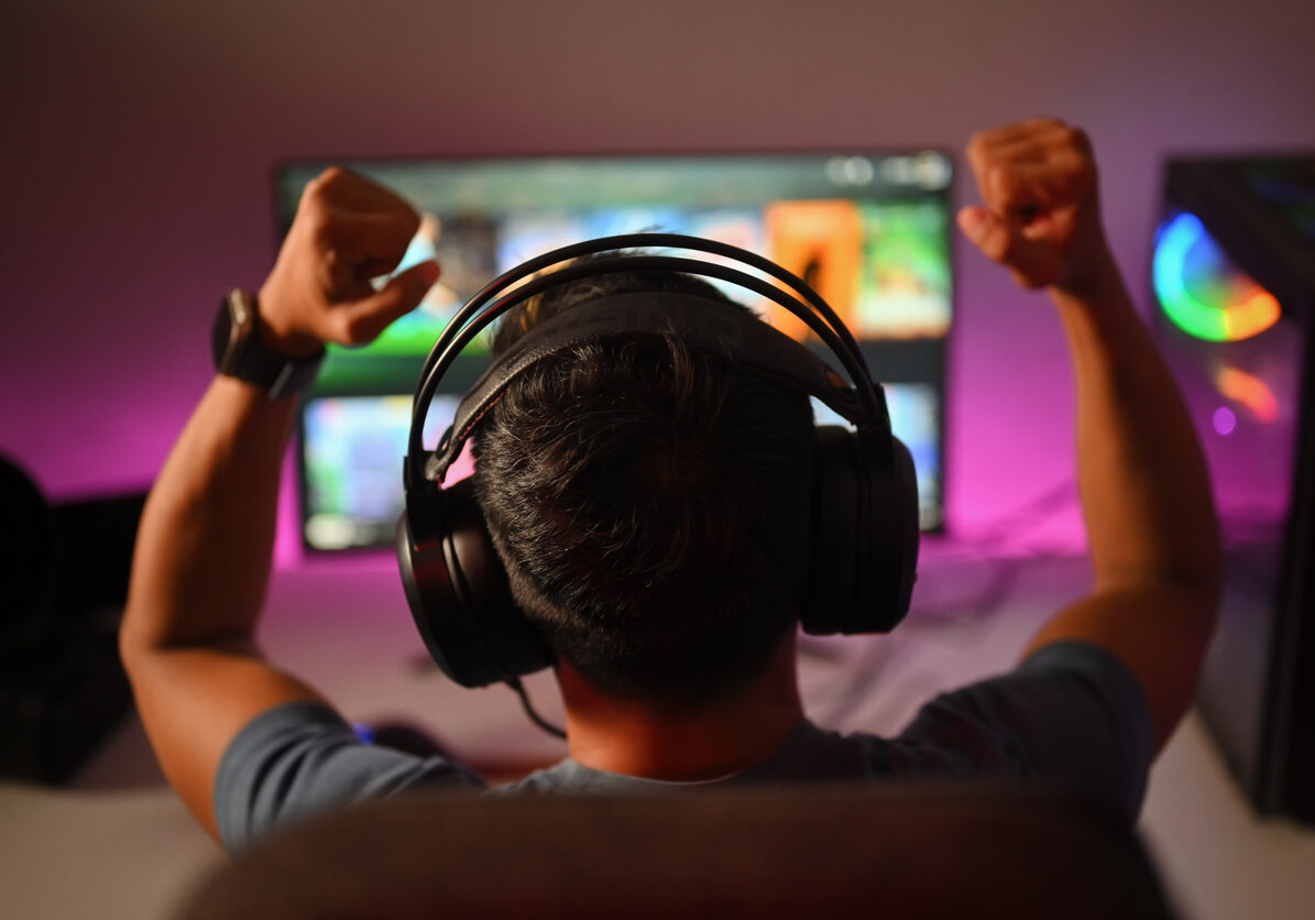 Back view of professional gamer wearing headphones celebrating victory while playing online video game at home.