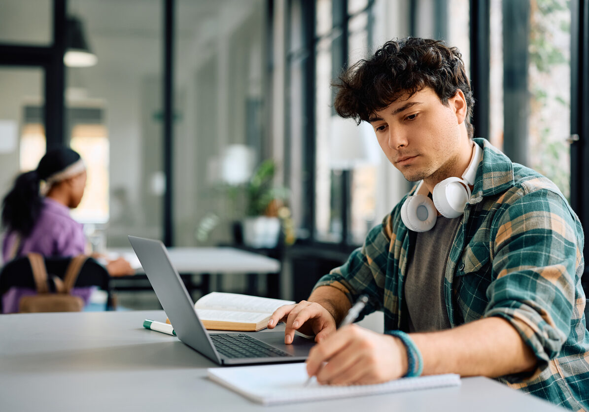 University student writing while using laptop and studying in the classroom.