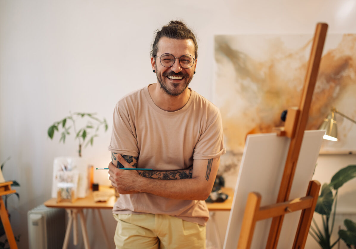 Young tattooed trendy artist standing at creative art studio with arms crossed and smiling at the camera. Portrait of confident professional painter standing at atelier and smiling at the camera.