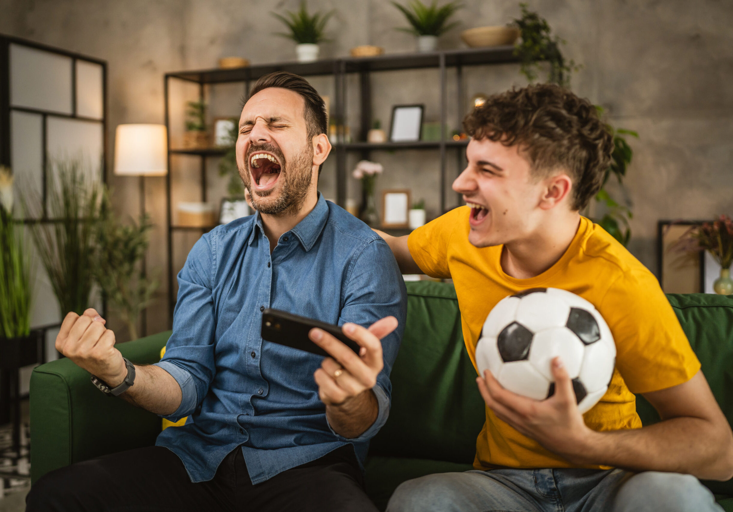 Two excited caucasian young adult men friends or brother use phone to watch football match and cheer at home