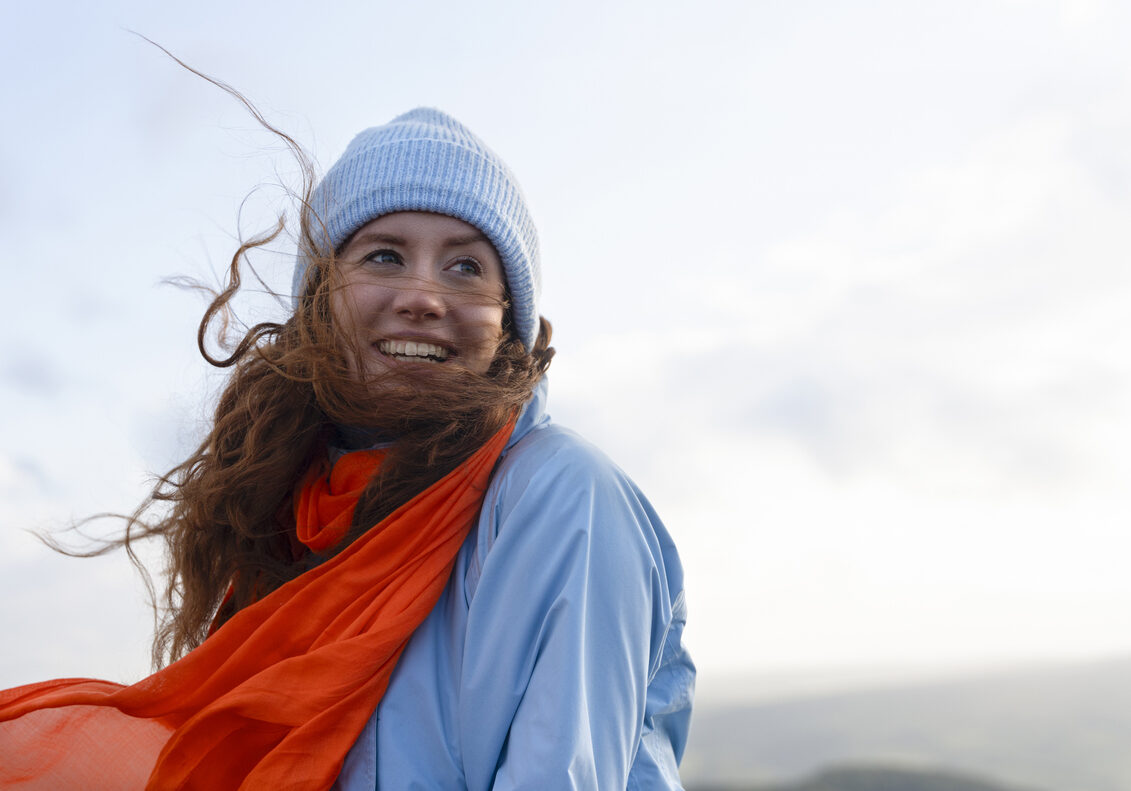 A young woman enjoys a windy day, smiling with her hair and scarf flowing in the breeze against a cloudy sky.