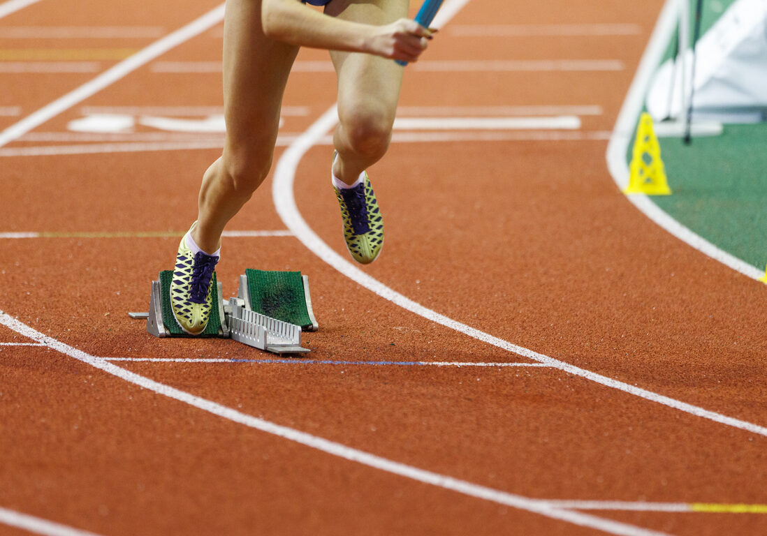 Athlete in sprinting shoes launches off the starting blocks during a competitive track race on an indoor arena surface.