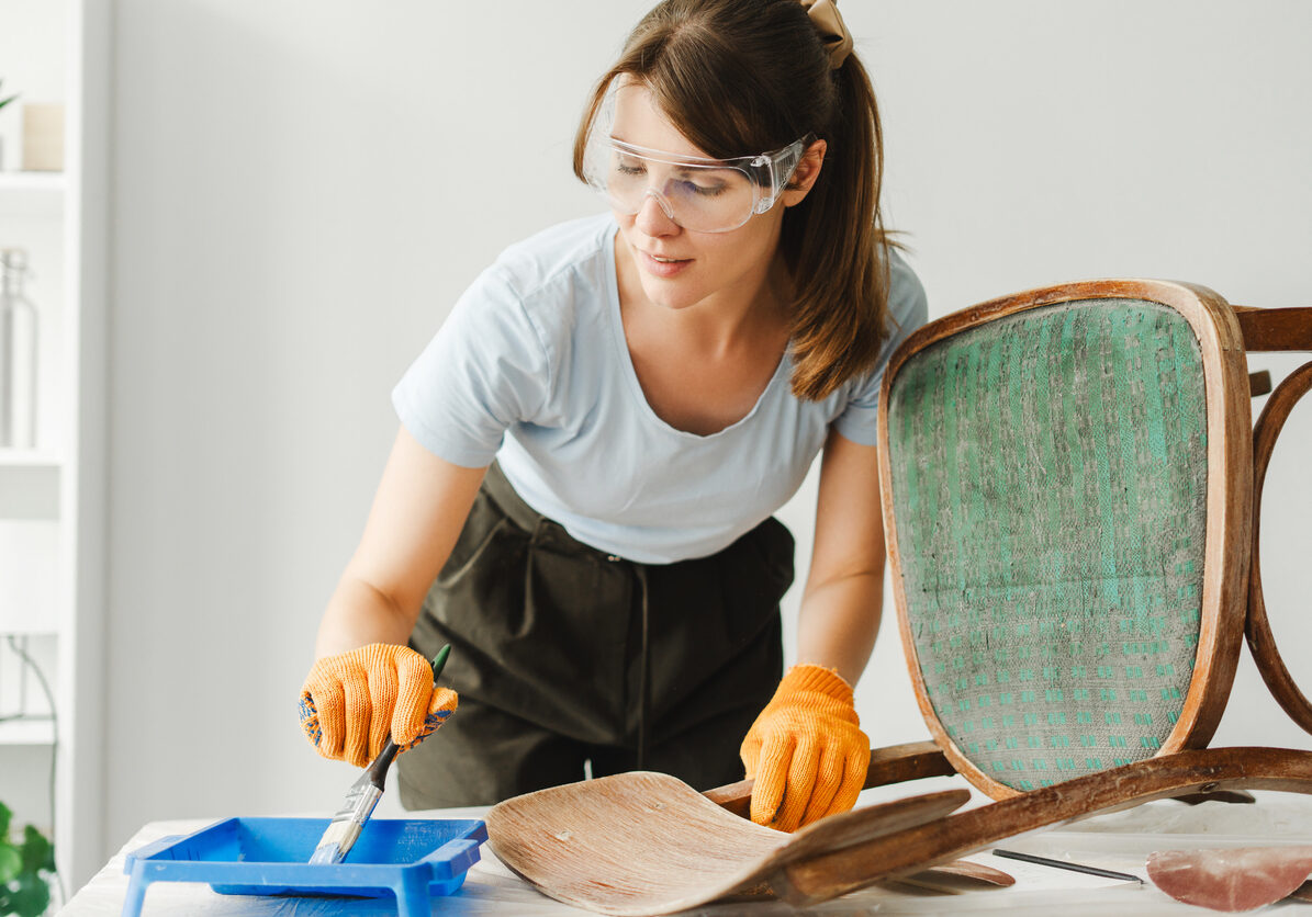 Craftswoman wearing protective gloves and glasses is restoring an old chair, carefully applying paint and varnish remover to its wooden parts