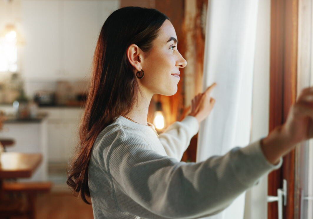 Woman, window and open curtains in house for start day, fresh air and check weather in living room. Girl, smile and drapes for awake, getting ready and morning routine with thinking of apartment view