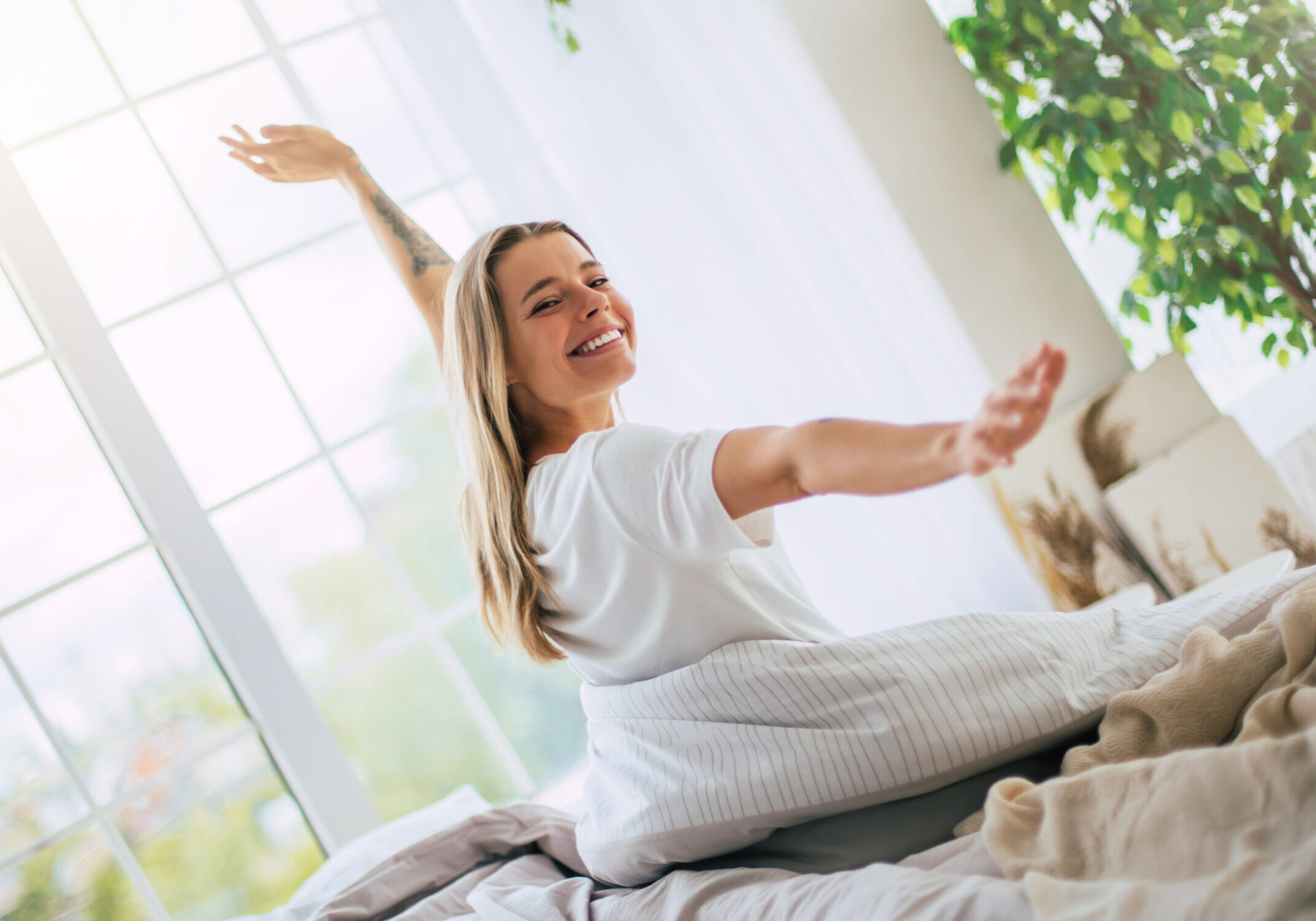 Cute smiling woman stretches joyfully on a bed, bathed in natural light from large windows. The bedroom is bright and cozy, with plants and soft bedding, evoking a sense of comfort and happiness