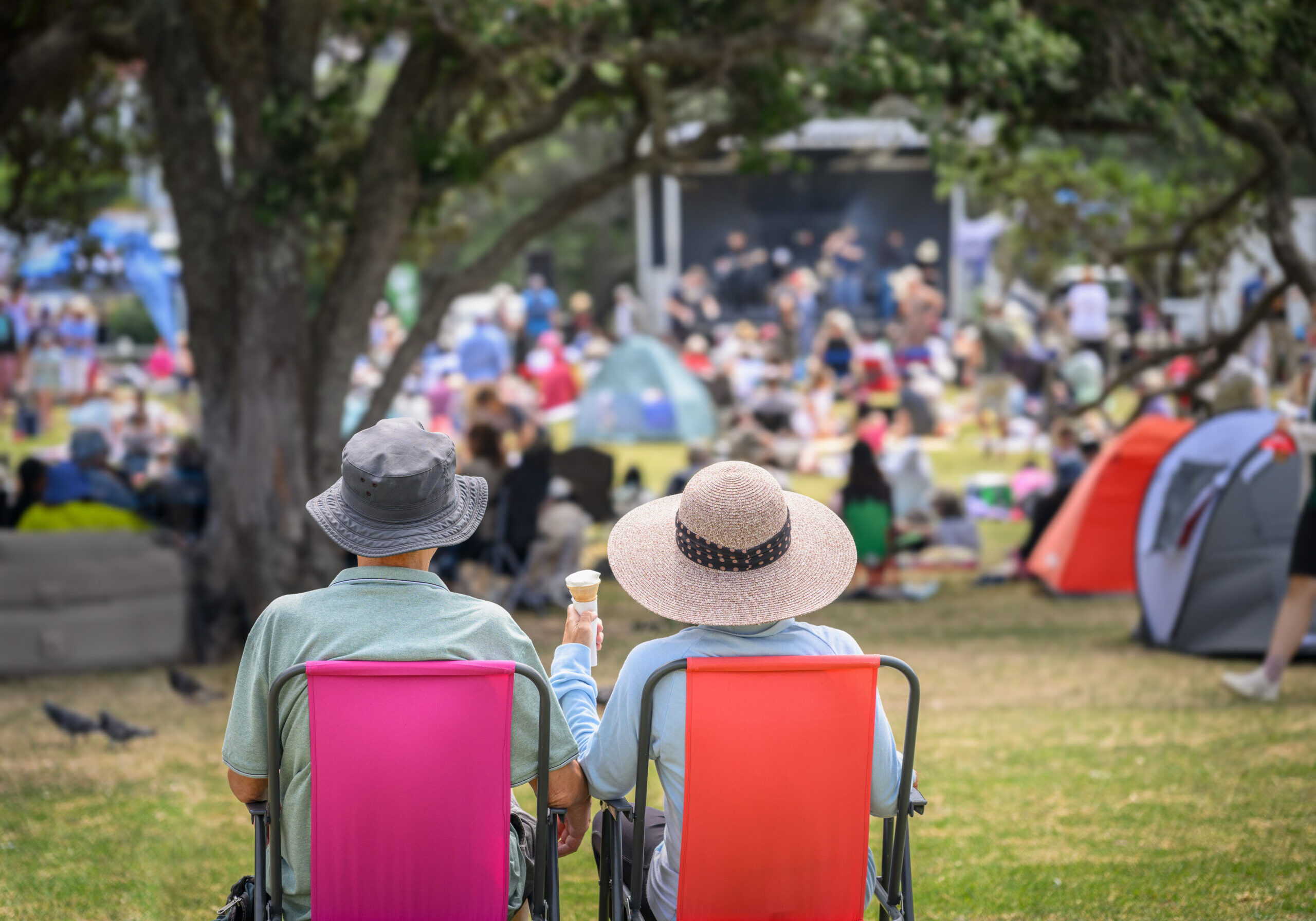 Couple sitting on camping chairs, unrecognizable crowds listen to the music concert in the park. Auckland.