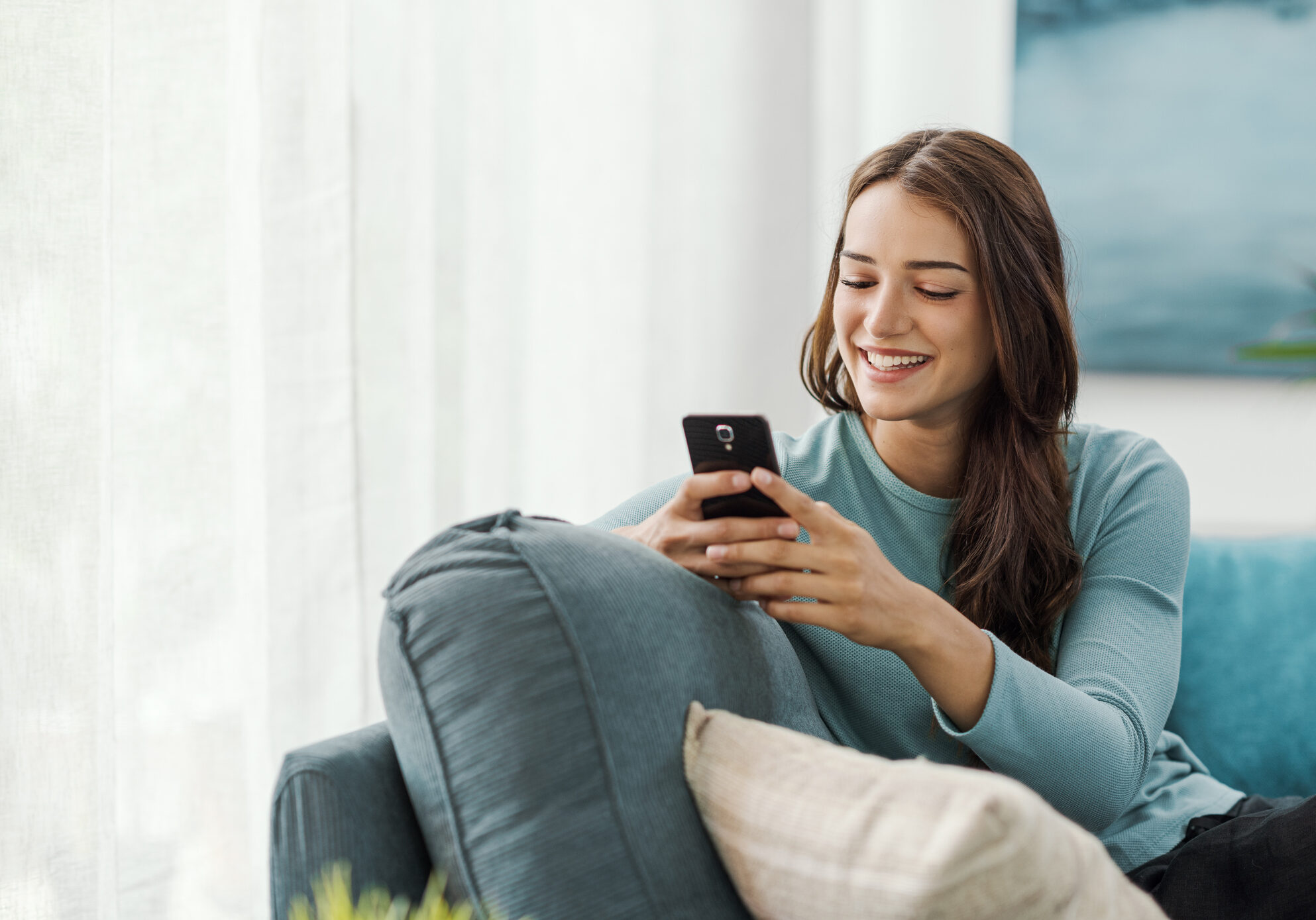 Happy young woman sitting on the couch at home and connecting with her smartphone, she is chatting and using mobile apps
