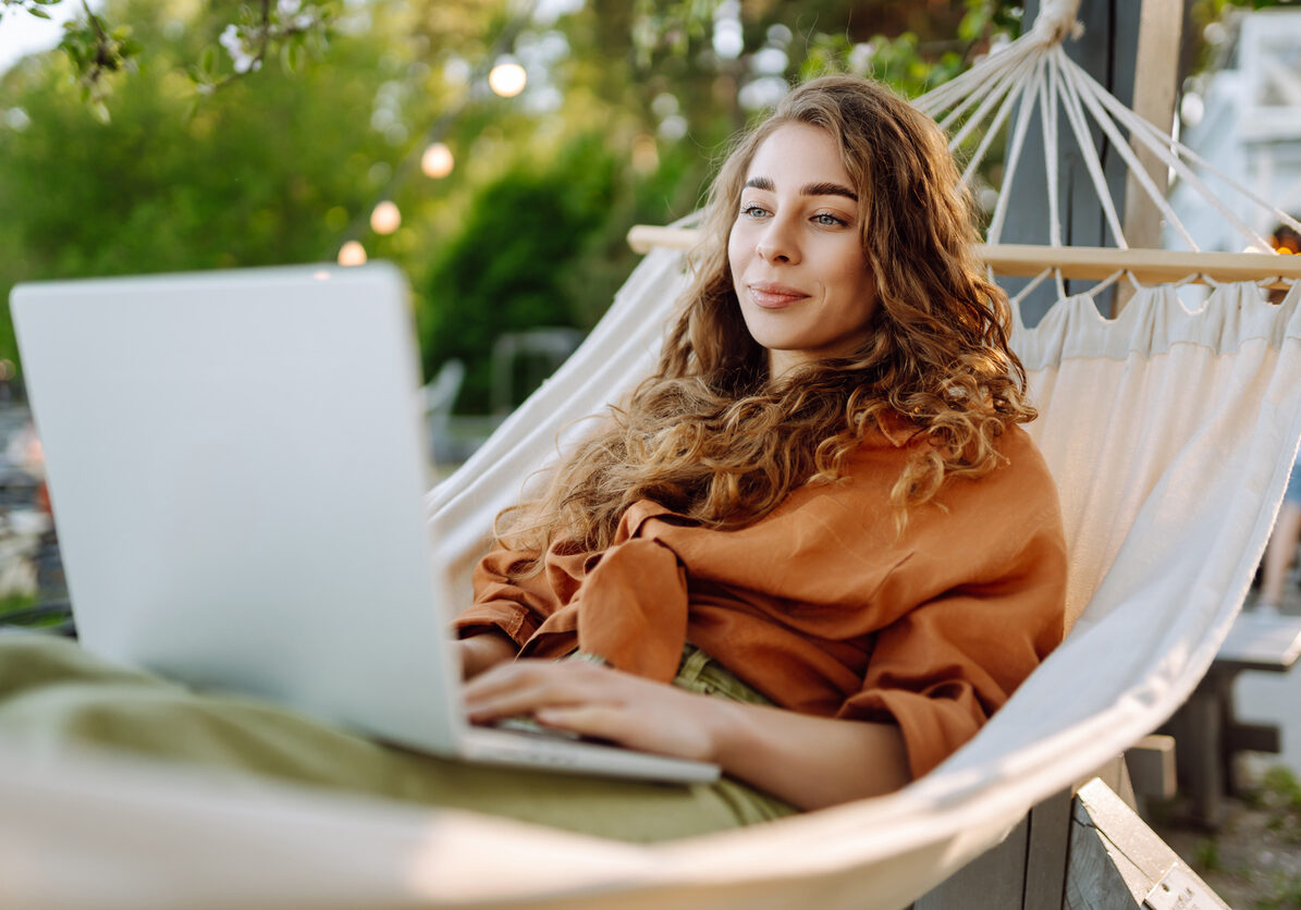 Young woman with laptop working lying in a hammock among trees in the fresh air. Female freelancer spending time in the park at sunset with laptop. Freelance, freedom concept. Active lifestyle.