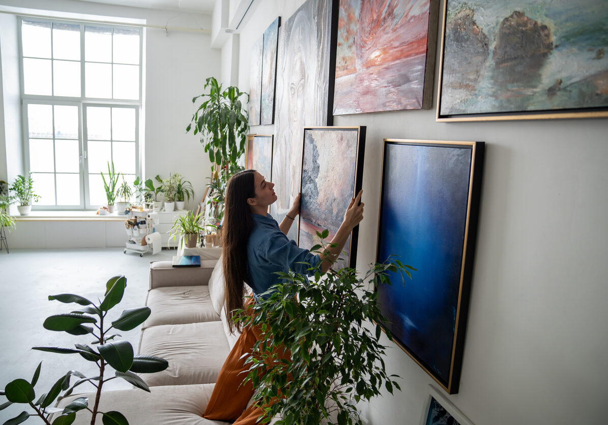 Woman painter hanging newly painted modern framed painting on wall in bright room art workshop with indoor plants. Actual solutions by decoration in contemporary interior design. Artists cosy home