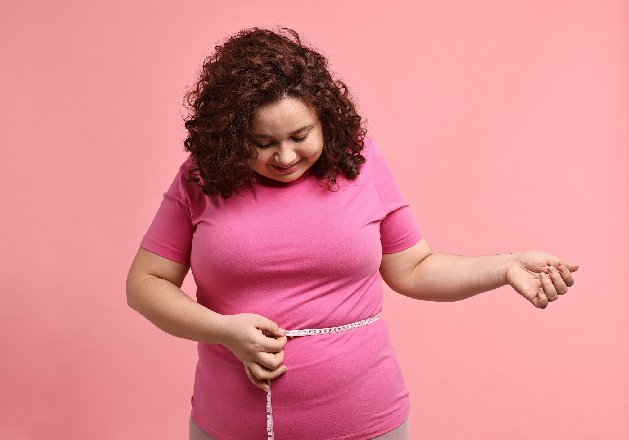 Weight loss. Happy plus size woman measuring waist with tape on pink background