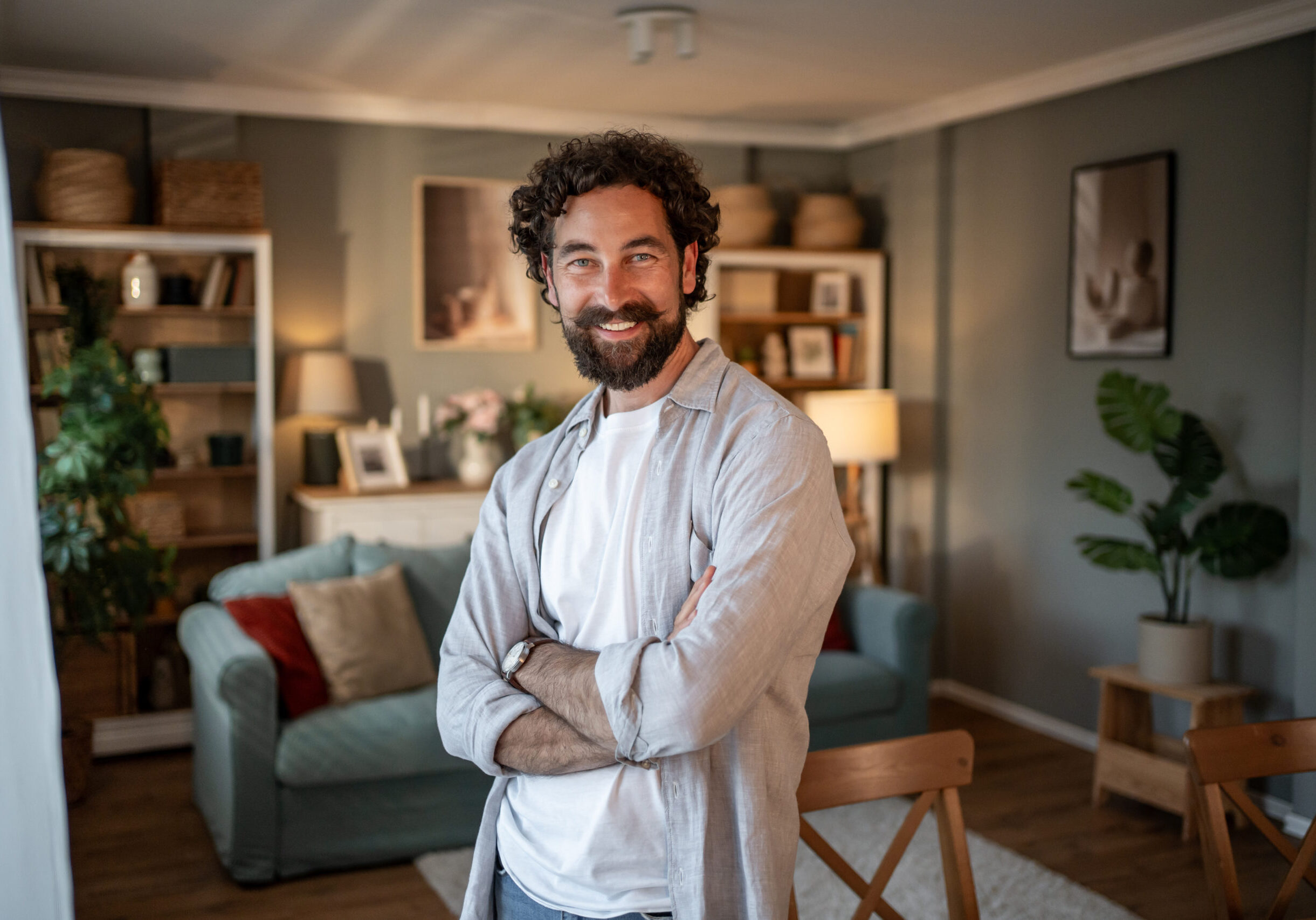 Mid adult man with curly hair and a beard is standing with his arms crossed, smiling at the camera in his modern, stylish apartment living room