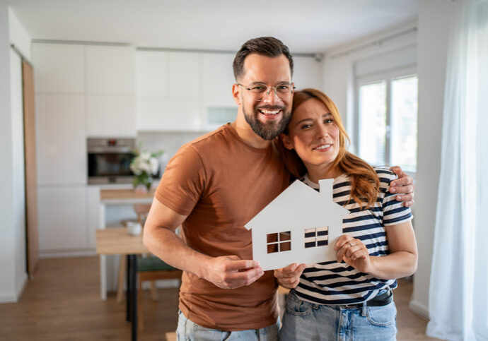Happy couple embracing and holding a white paper house, smiling at the camera in their modern new home, celebrating buying their first house together