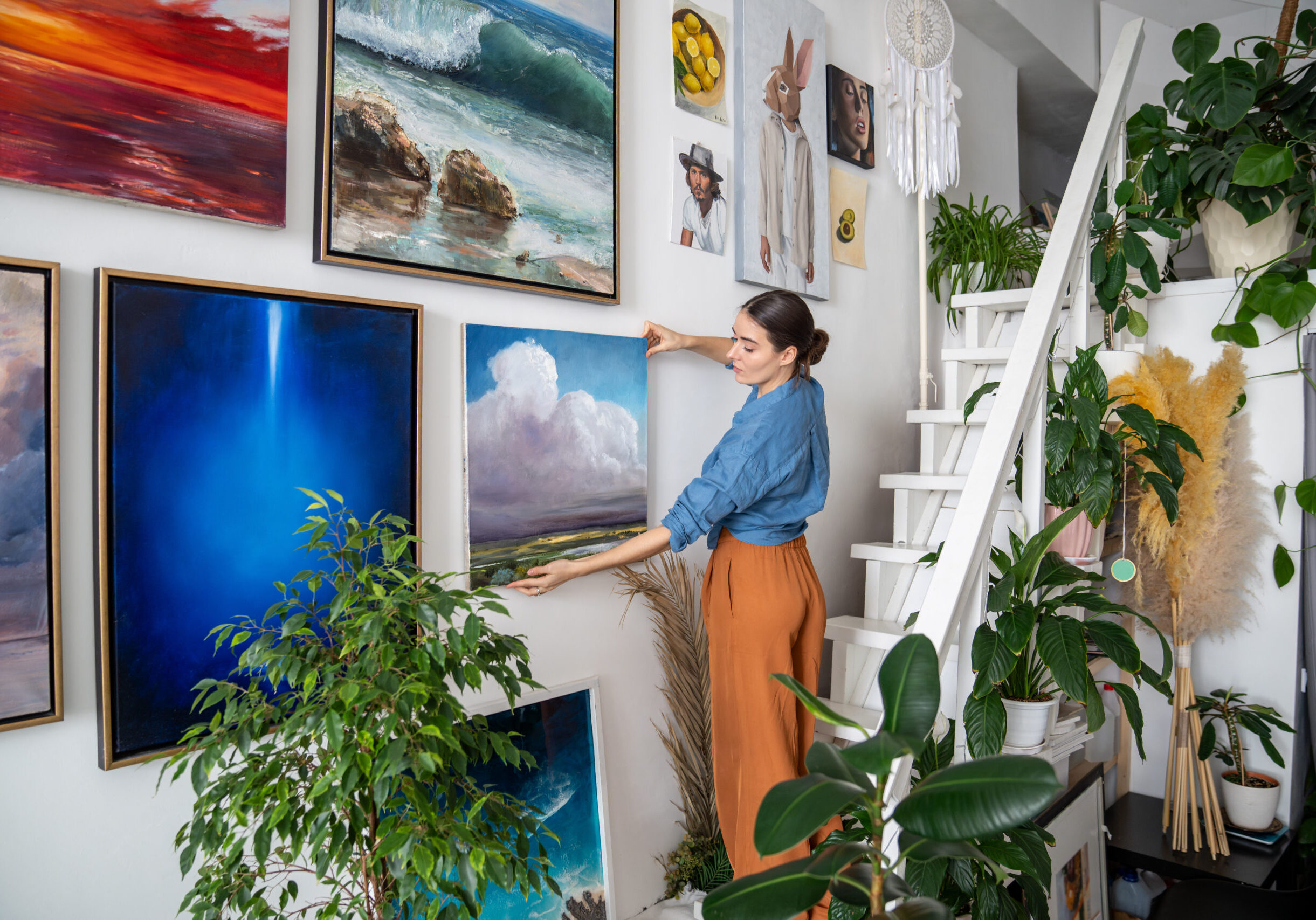 Serious woman hangs pictures on wall. Concentrated unsmiling female artist creates composition of paintings, decorations on wall. Creative designer in art home studio workshop among artworks plants.