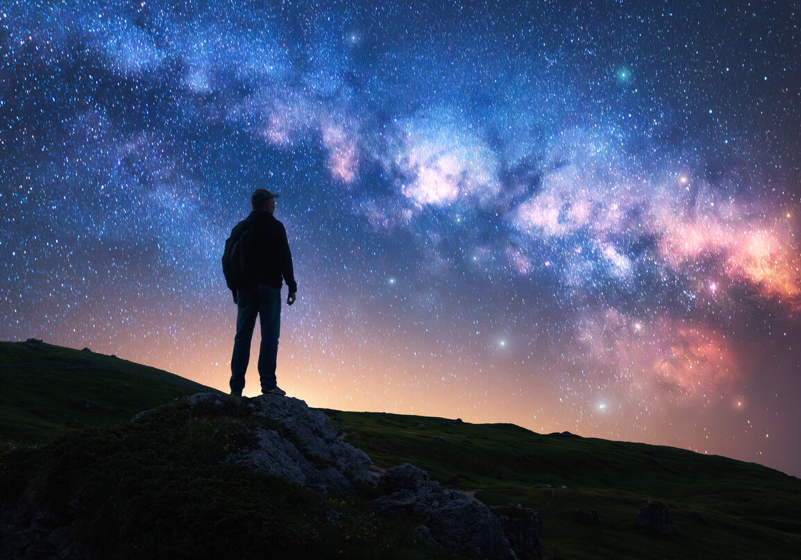 Glowing Milky Way and man on mountain trail at starry night. Silhouette of a guy on the hill, sky with bright stars. Galaxy. Space landscape with milky way. Nightscape. Travel background. Concept