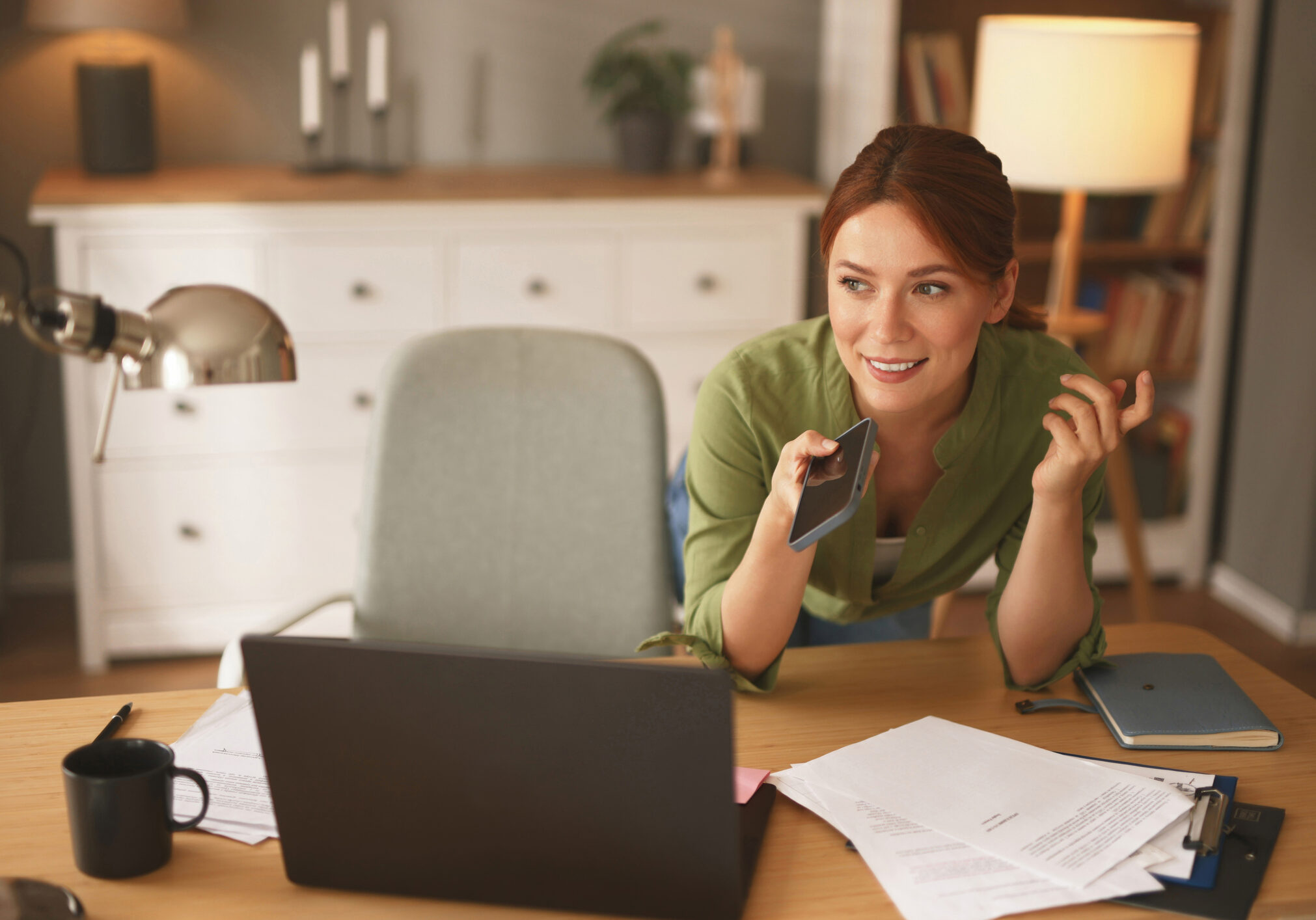 Smiling woman recording voice message on smartphone and working on laptop at home