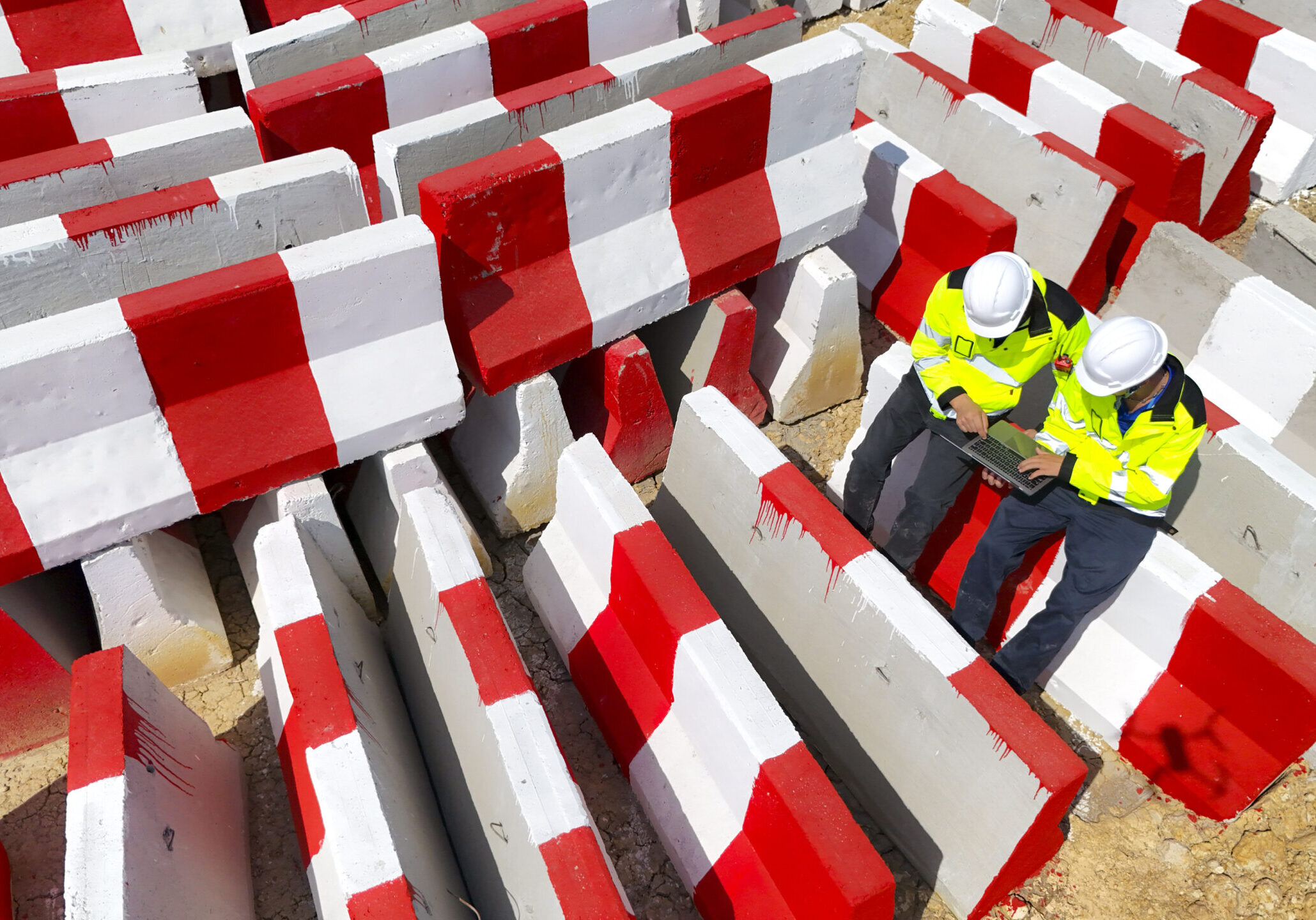 Top view of two construction workers in safety gear using a laptop at a worksite surrounded by red and white barricades, showcasing teamwork, planning, and efficient use of technology in construction