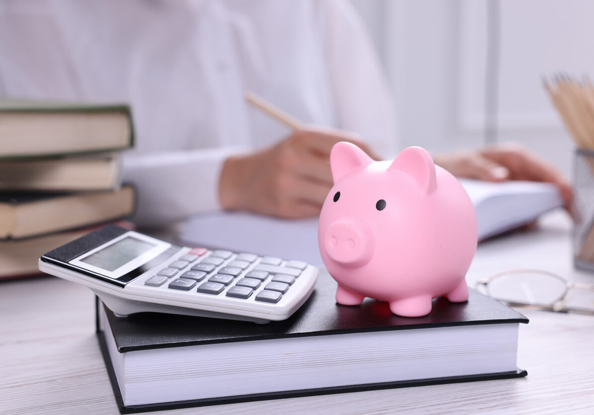 Piggy bank with calculator, book on white wooden table and woman working indoors, selective focus