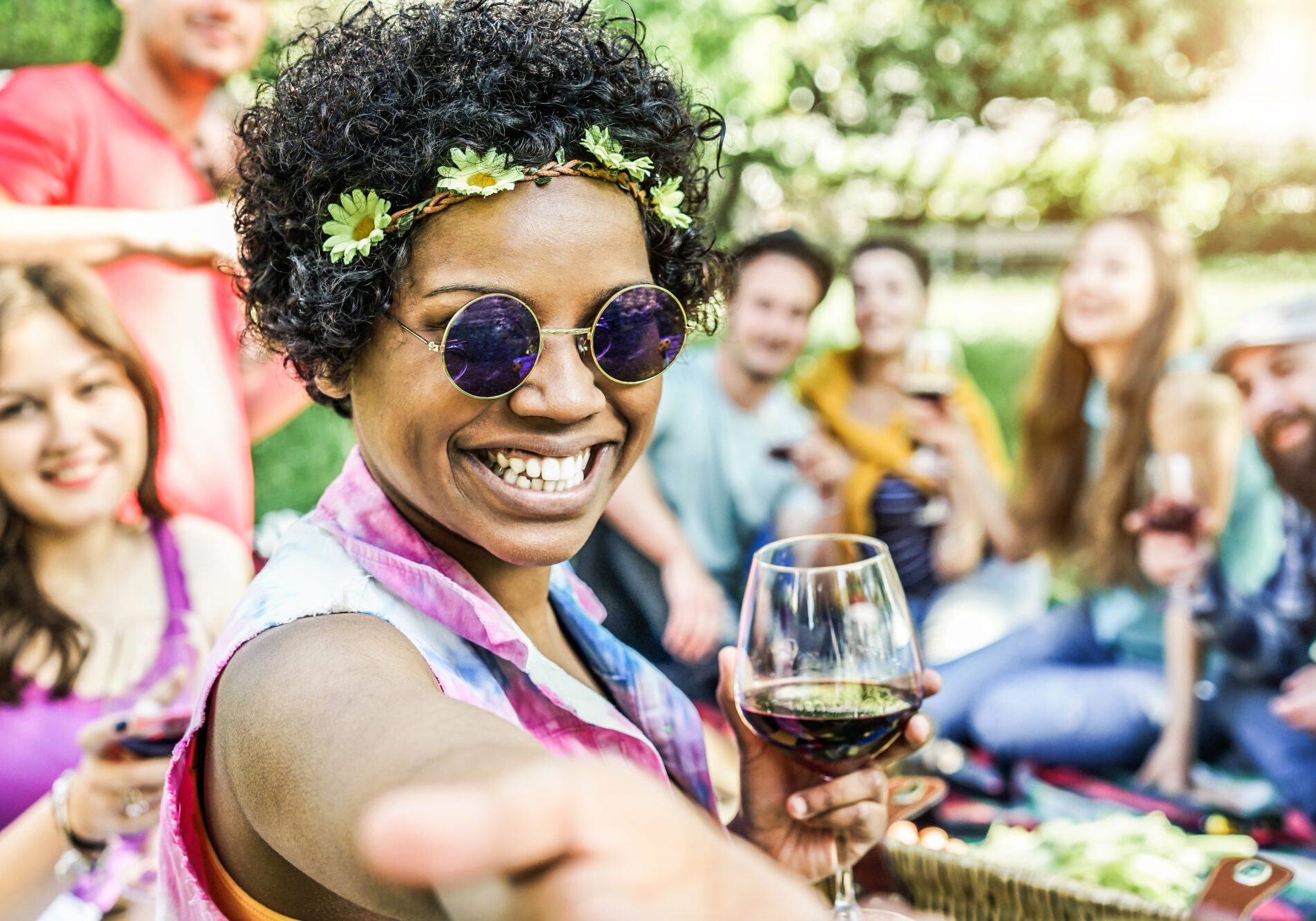 Happy mixed race woman taking selfie at picnic outdoor with her friends - Young trendy people having fun while eating and toasting red wine - Youth and friendship concept - Warm contrast filter