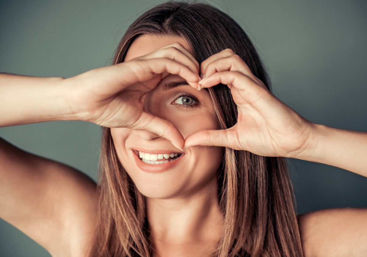 Beautiful woman is showing heart, looking at camera and smiling, on gray background