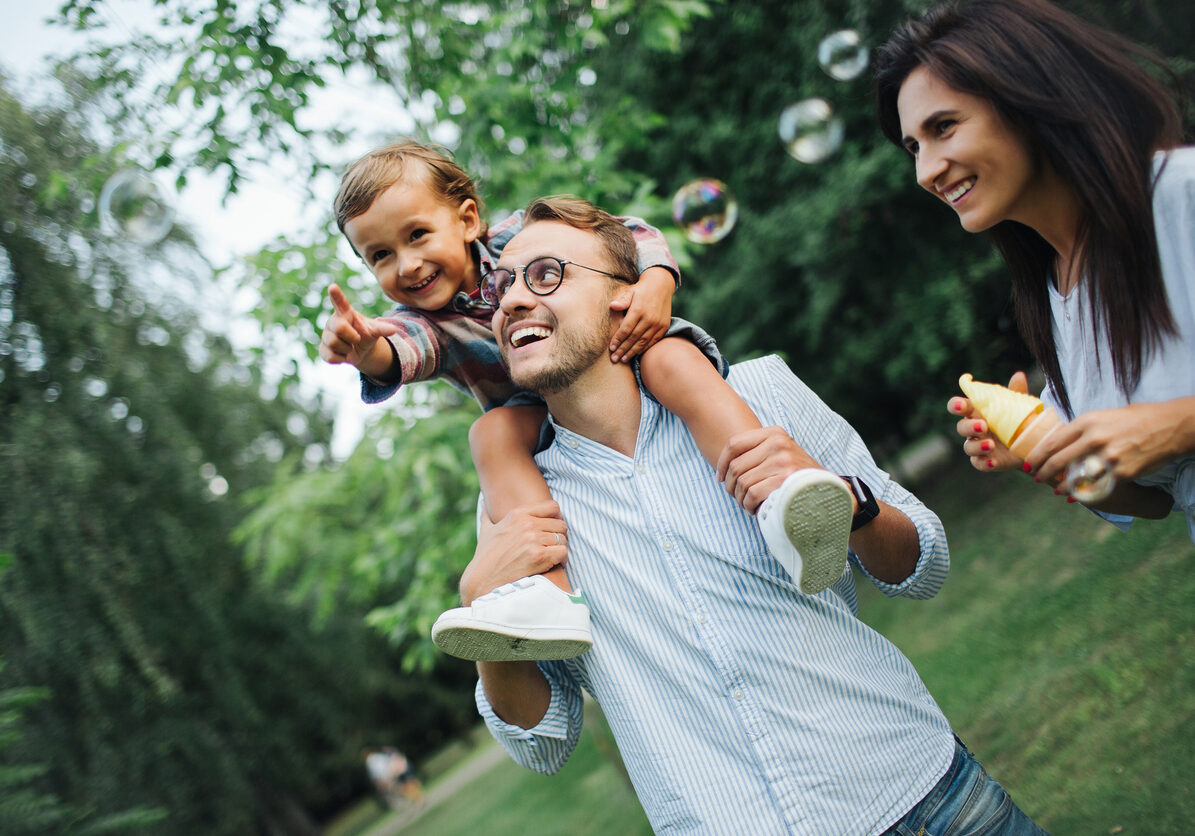 Happy young family playing with bubble wands with son sitting on fathers shoulders in park outdoors in summer