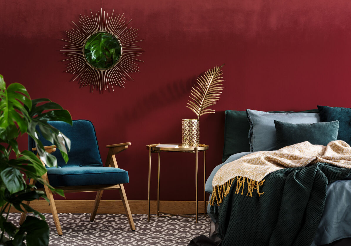 Gold table between green armchair and bed in sophisticated red bedroom interior with mirror