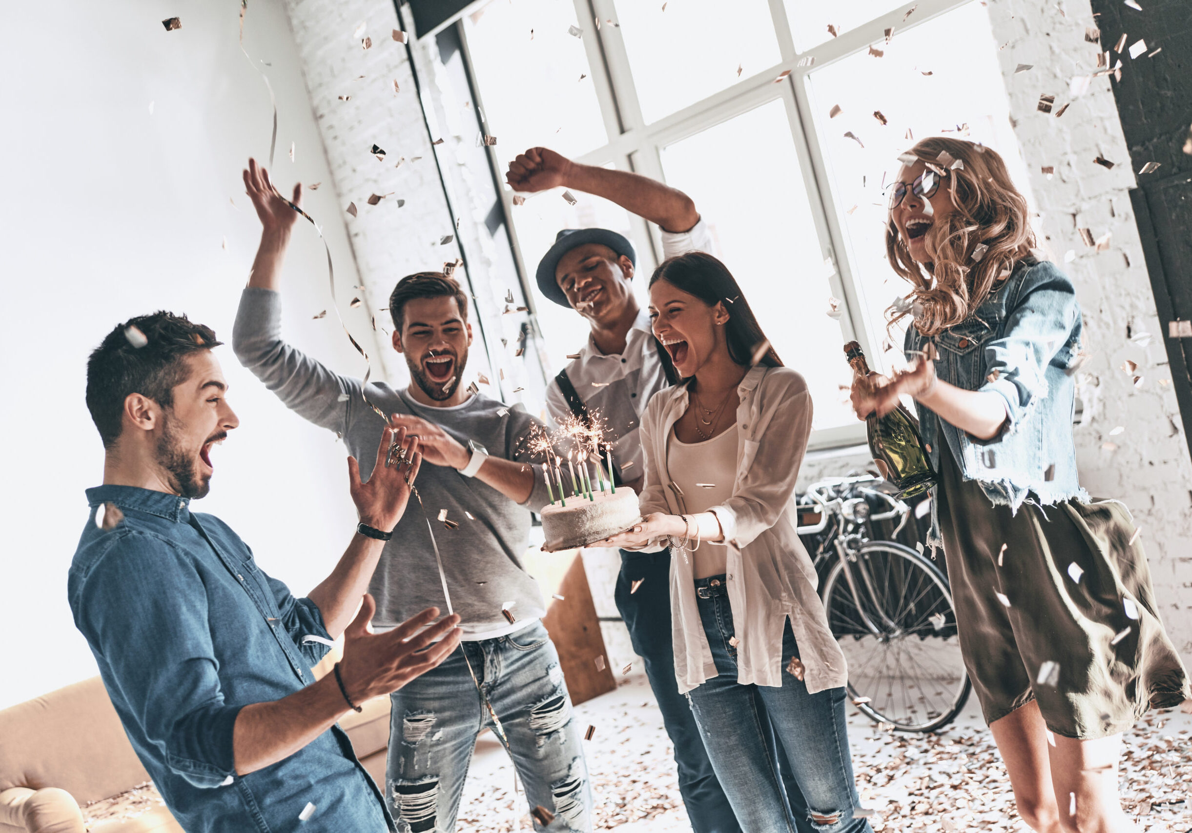 Happy young man celebrating birthday among friends while standing in room with confetti flying around
